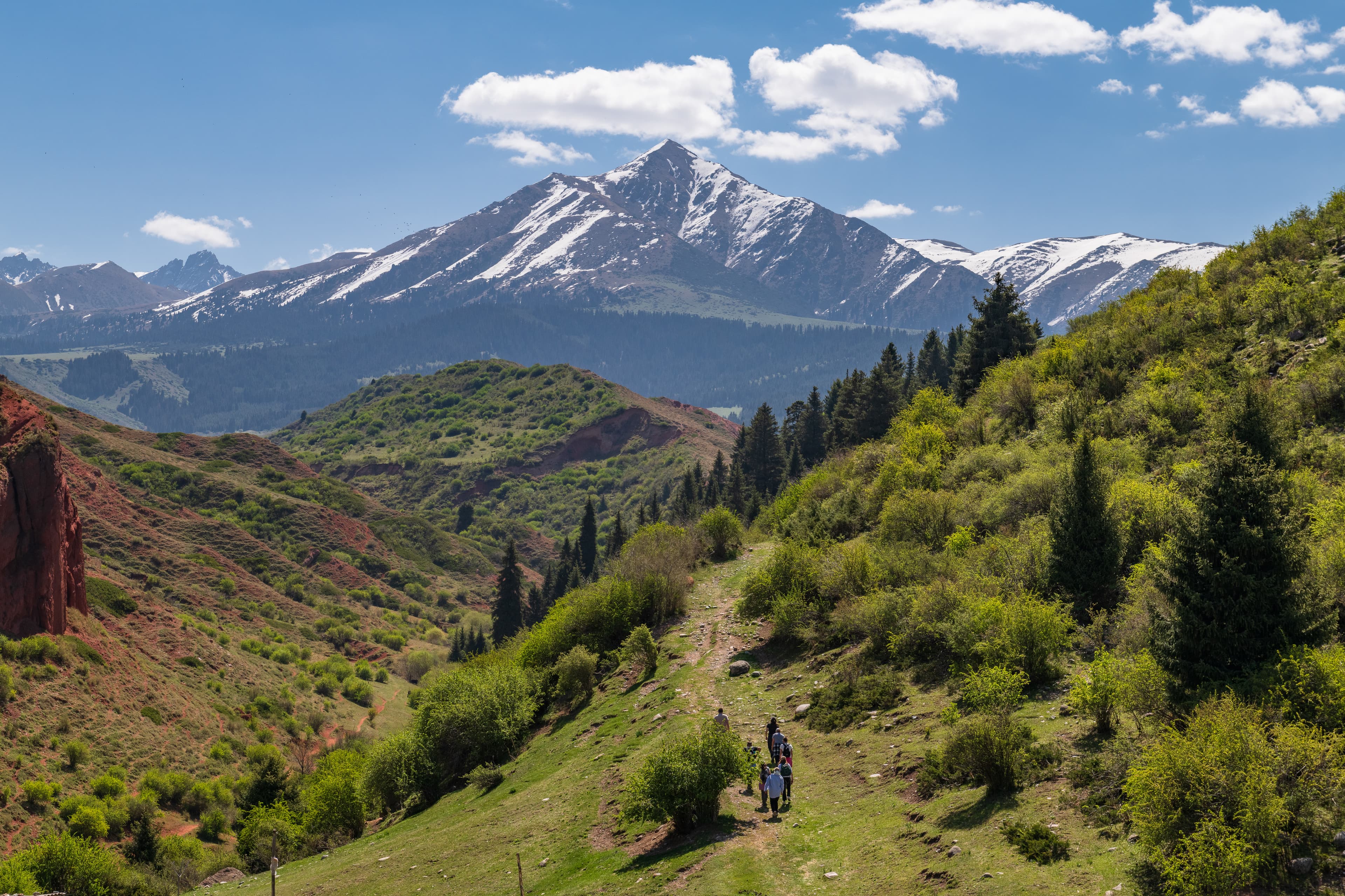 The snowy Jeti Oguz peak stand in the middle above red canyons and green juniper and fir-tree forests